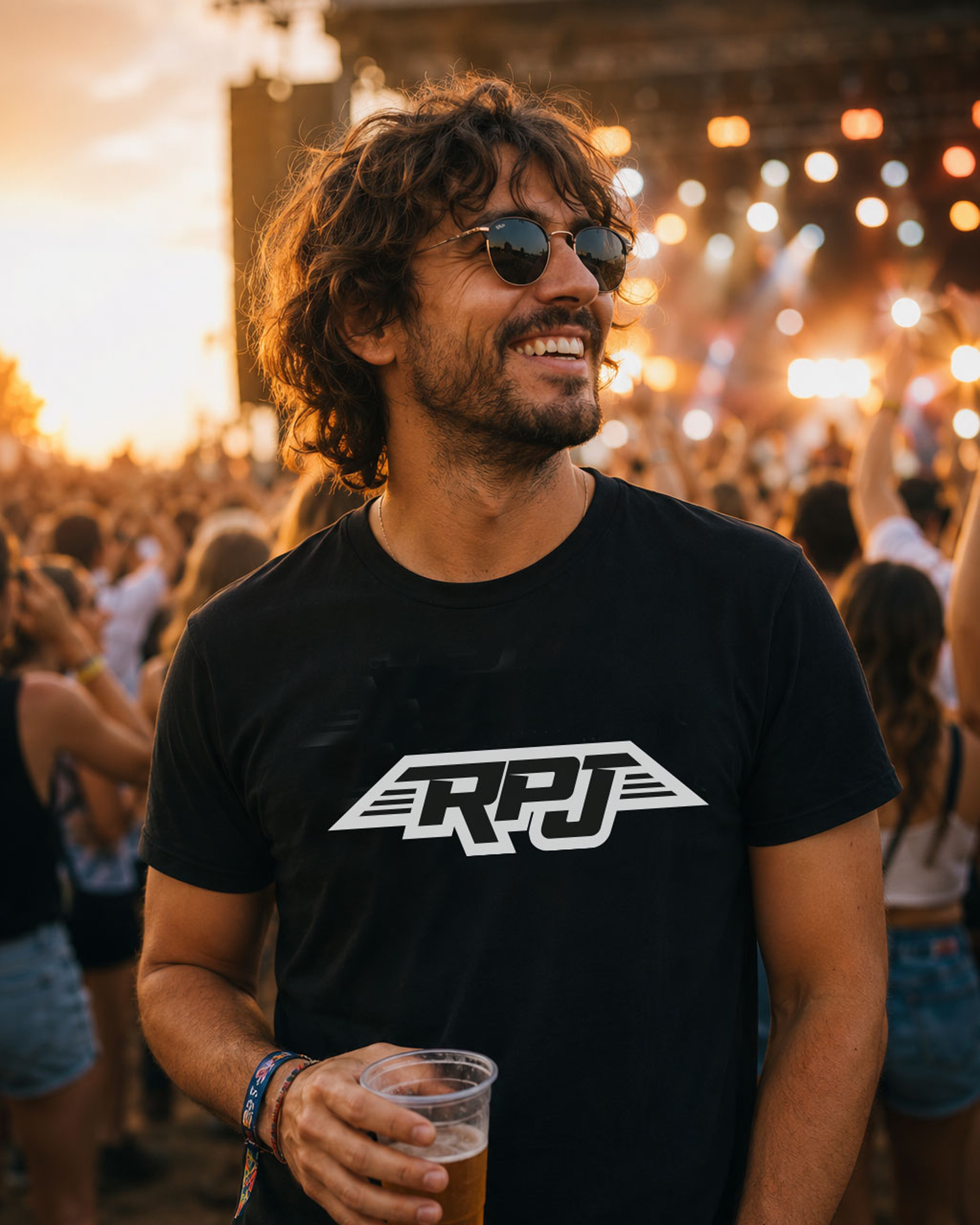 A smiling man with curly hair and sunglasses at an outdoor concert or festival, wearing a black RPJ brand t-shirt and holding a plastic cup of beer.