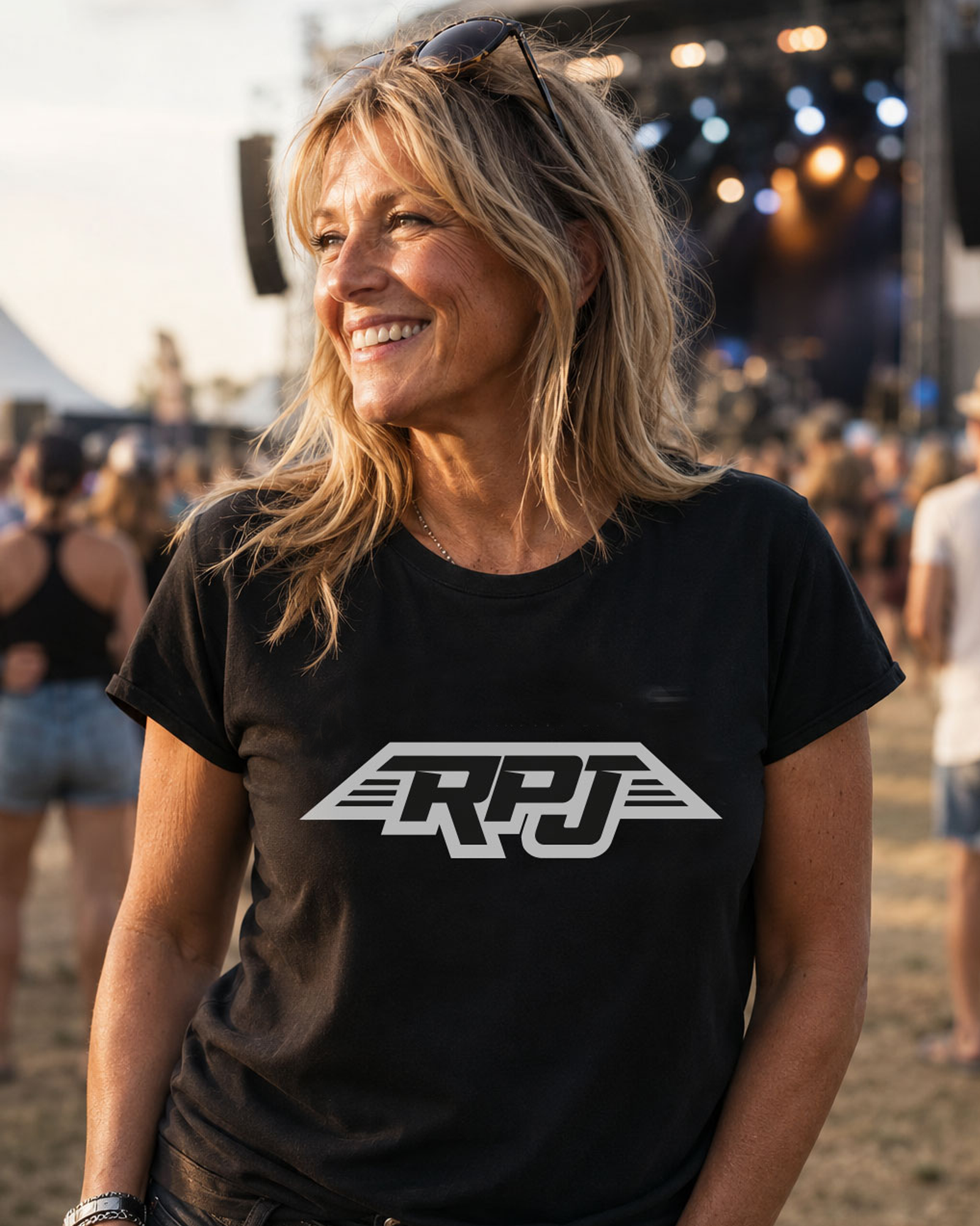 A woman smiling outdoors at a festival or concert, wearing a black short-sleeve T-shirt with a large white stylized 'RPJ' logo across the chest.

